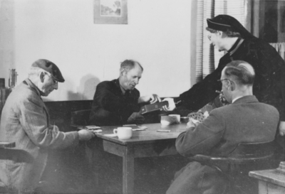 876595 Portret van drie mannen die aan het kaarten zijn en een Zuster Augustines die een koekje aanbiedt, tijdens een ...
